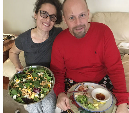 Paul and Jomania are sitting on the couch and smiling at us.  Jomania is holding a large bowl with salad and Paul is holding a plate with his dinner partly eaten.